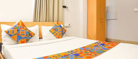 A hotel room with a bed, two pillows with geometric patterns, a wooden wardrobe, and a window with curtains.