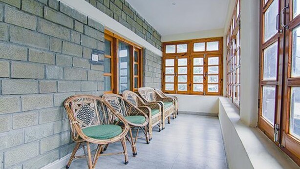 A corridor with wicker chairs lined up against a brick wall, large windows on one side, and a wooden door on the other. - Fabhotel Arigato Cottage (Manali)