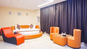Une chambre d’hôtel moderne avec un lit aux lignes courbes, des fauteuils orange, une petite table et une grande fenêtre avec des rideaux.