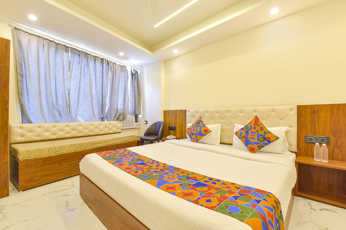 A hotel room with a large bed, a sofa bed, a wooden headboard, a bedside table, and a window with curtains.