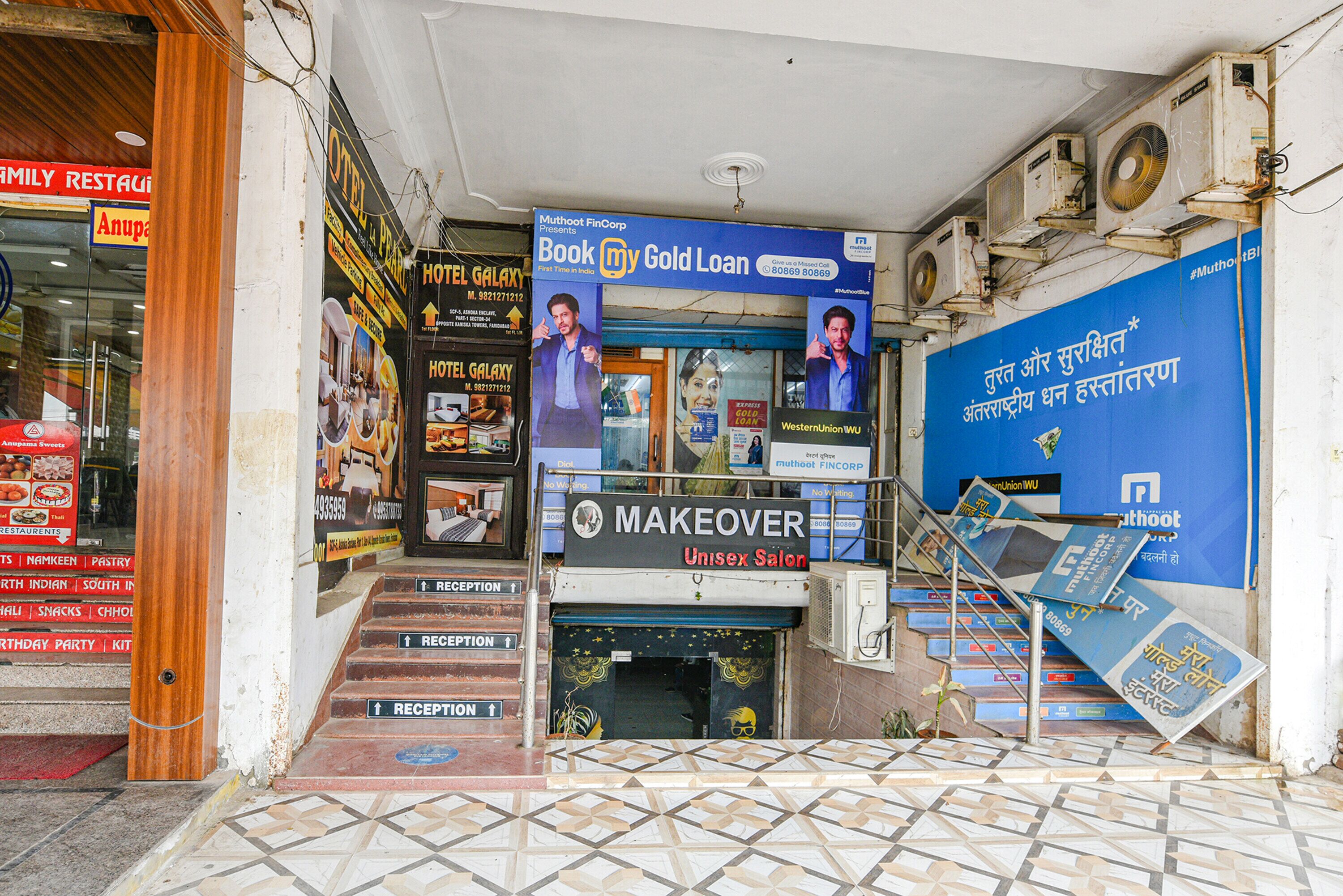 A hotel entrance with a reception desk, a staircase, and multiple signs including a 'Makeover Unisex Salon' and a 'Hotel Galaxy' sign.