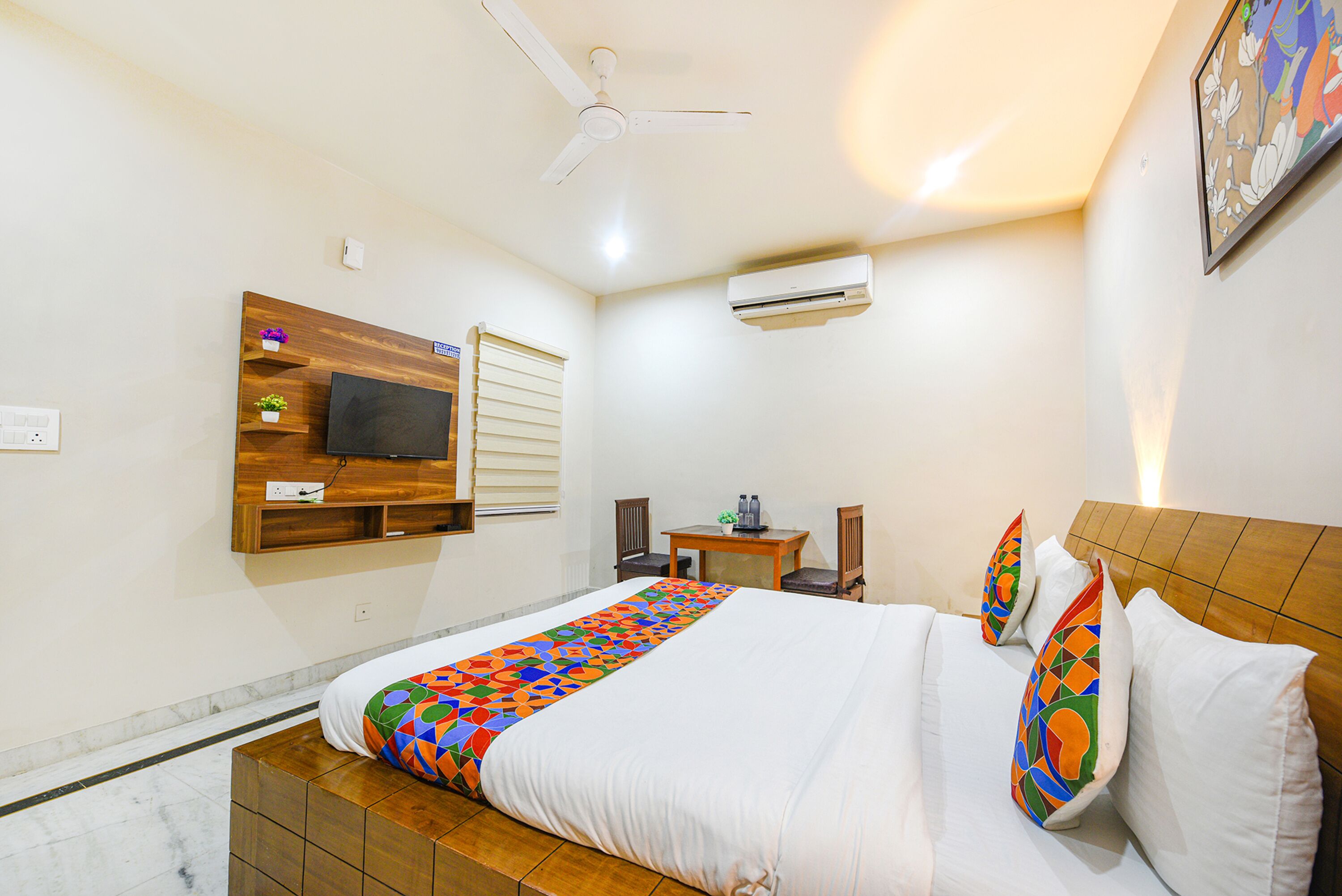A hotel room with a bed, a small table, a chair, a TV, and an air conditioning unit.