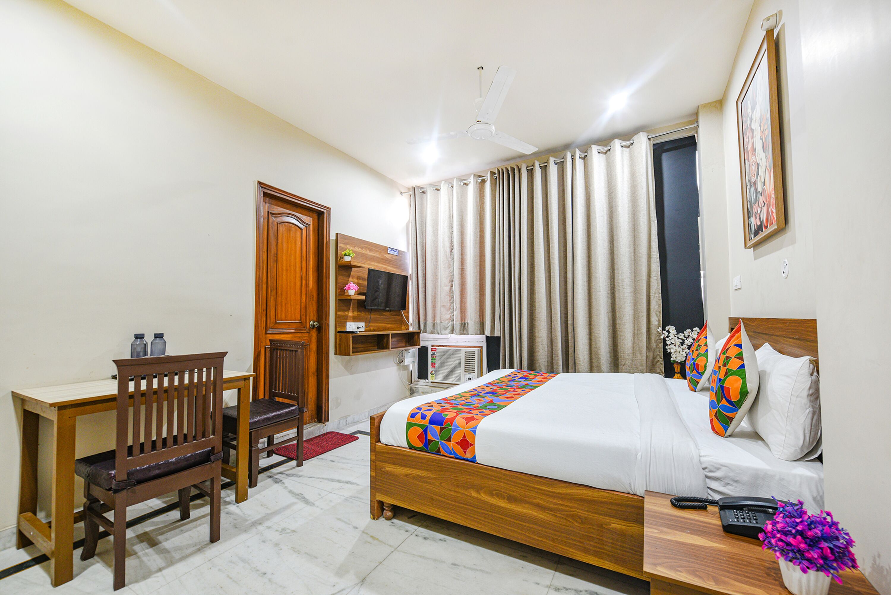 A hotel room with a wooden bed, a desk with a chair, a ceiling fan, and a window with curtains.