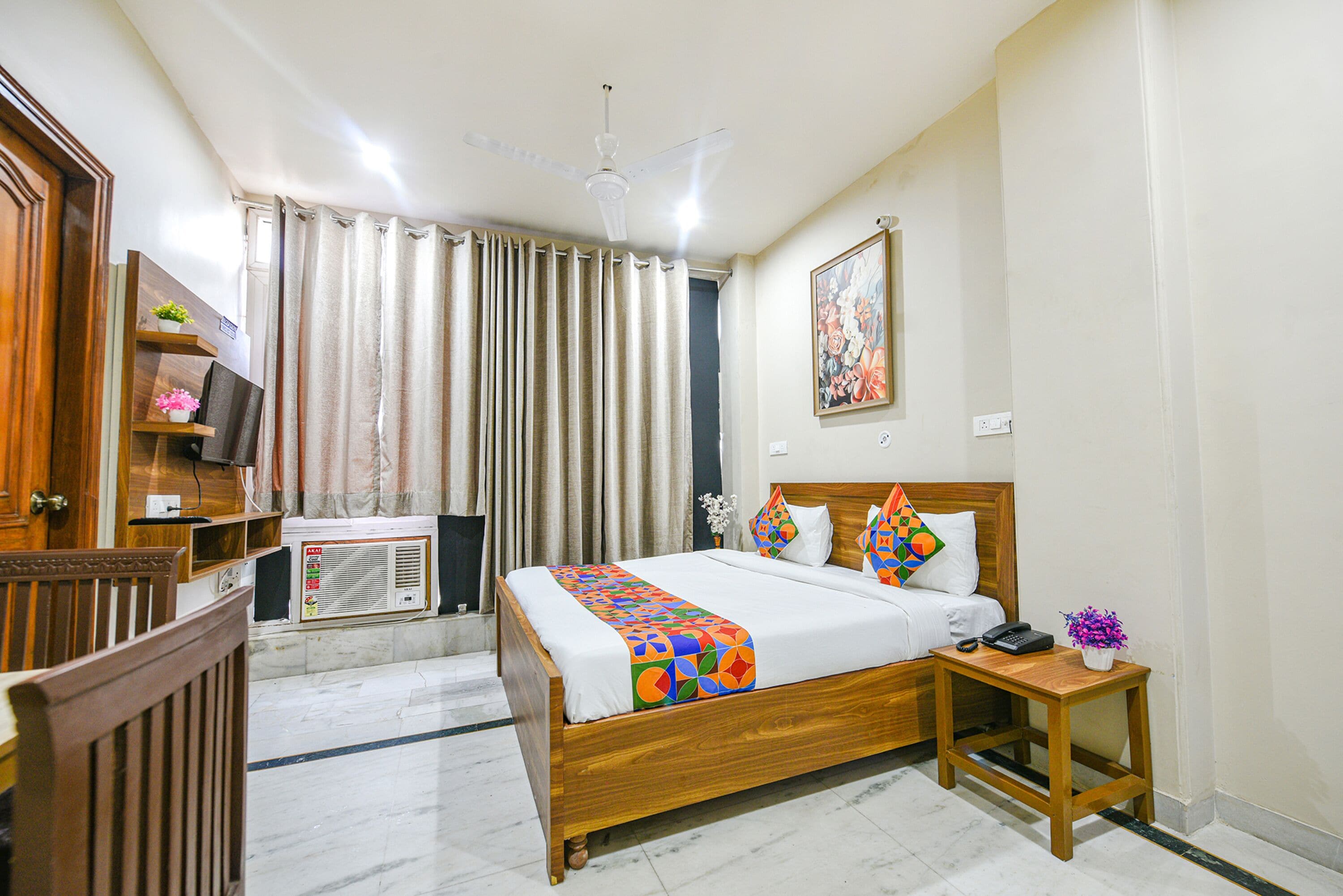 A hotel room with a wooden bed, a bedside table with a phone and a vase of flowers, a ceiling fan, and a window with curtains.