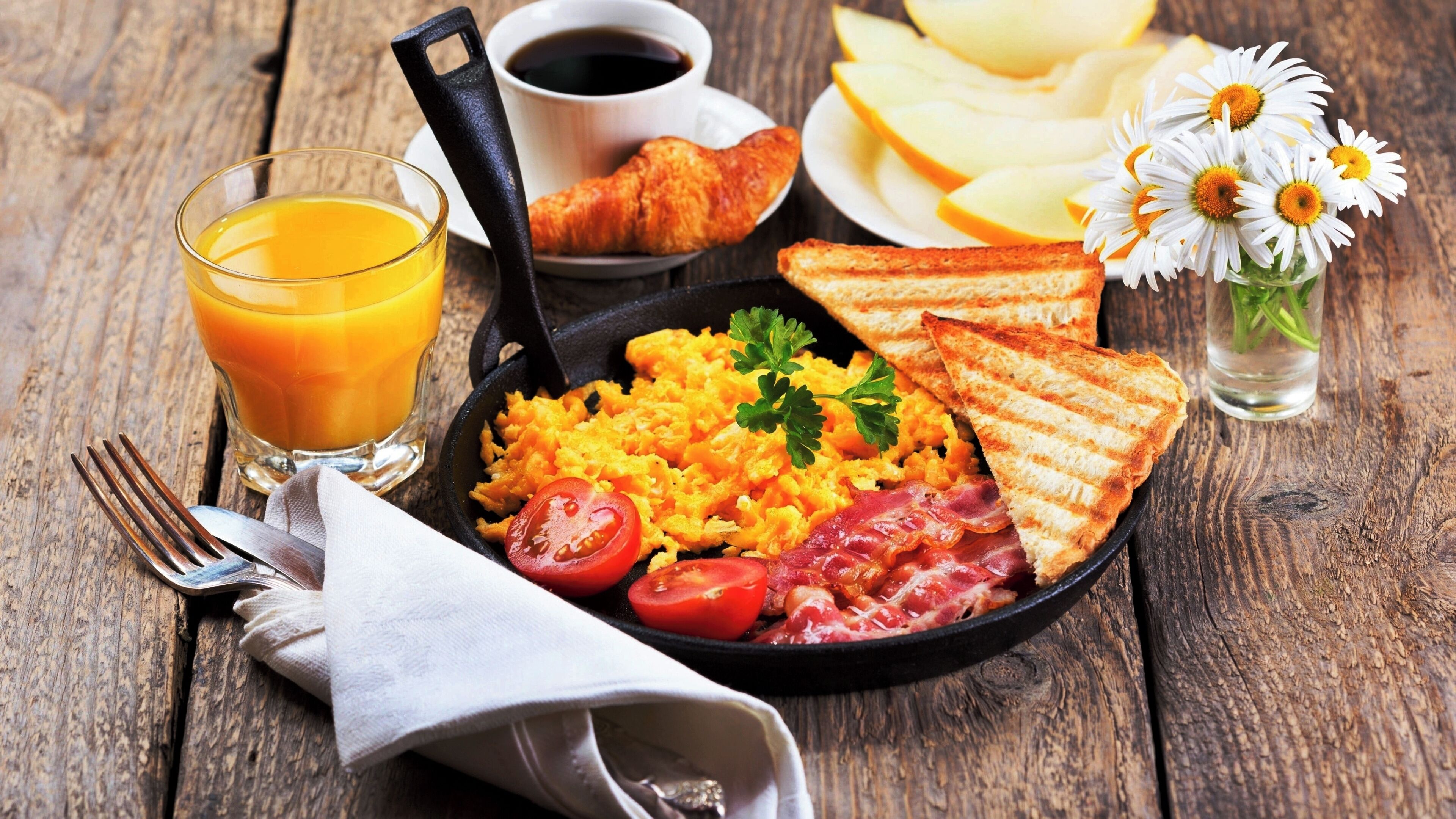 Ein Frühstücksteller mit Rührei, Speck, Tomaten und Toast, dazu ein Glas Orangensaft und eine Tasse Kaffee.