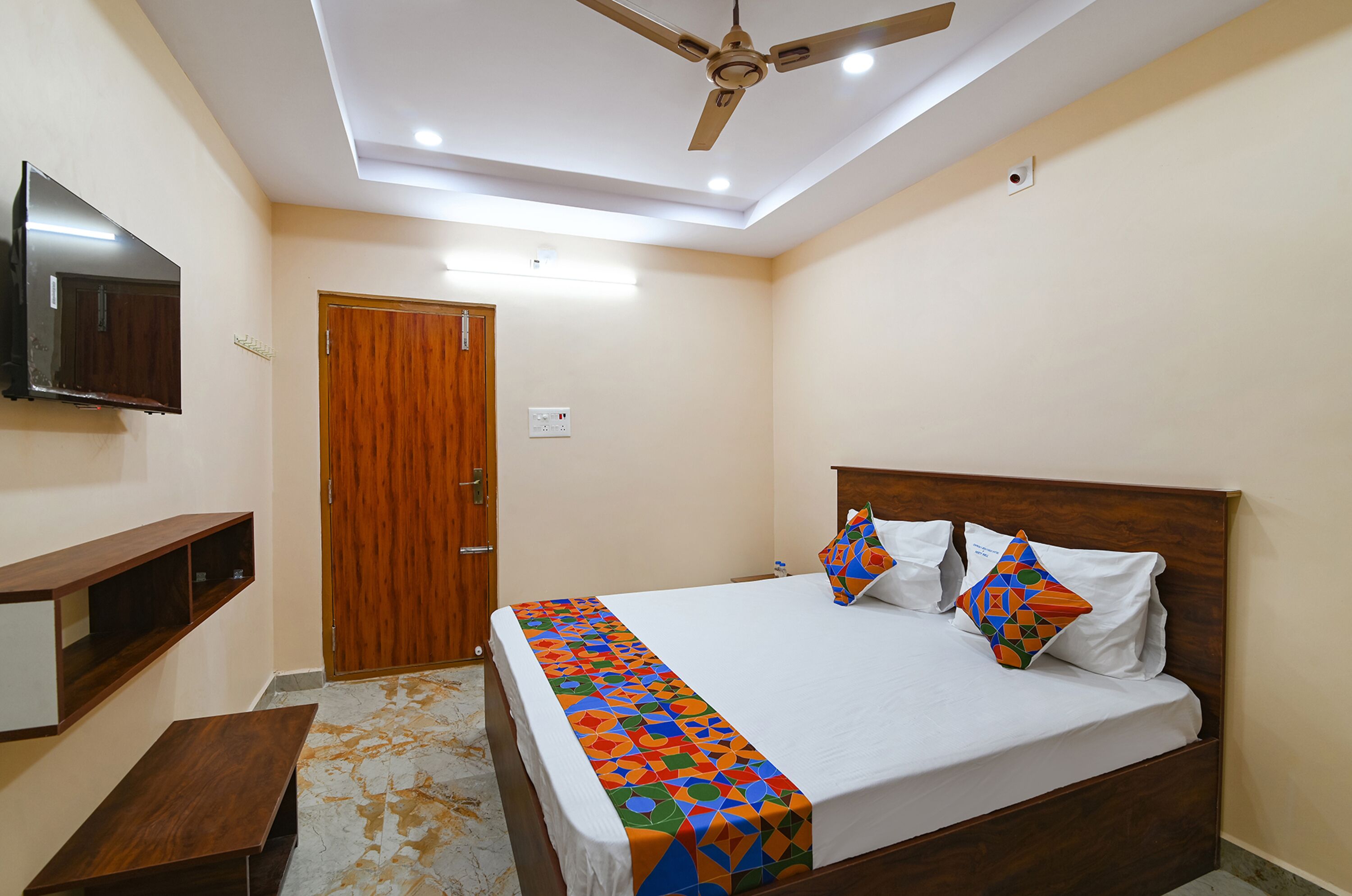 A bedroom with a bed, a ceiling fan, a TV mounted on the wall, and a wooden door.