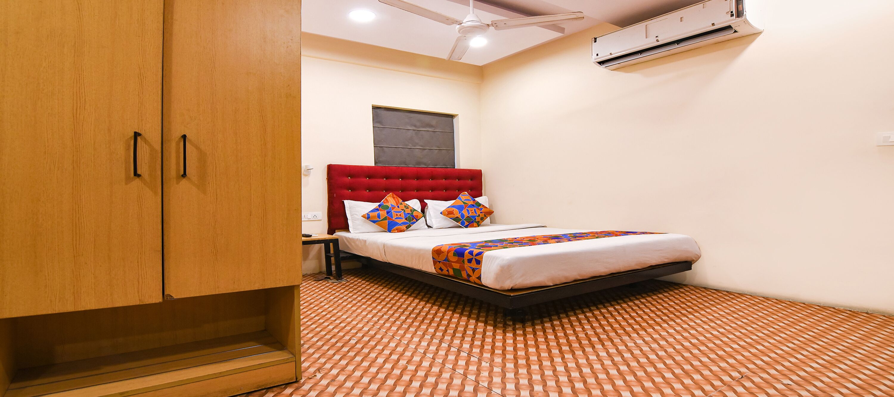 A bedroom with a red tufted headboard, a bed with a colorful blanket, a wooden wardrobe, and a ceiling fan.