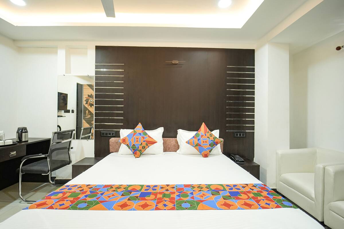 Habitación de hotel con una cama grande, dos almohadones decorativos, un escritorio con silla, una mesita de noche y un sillón blanco.