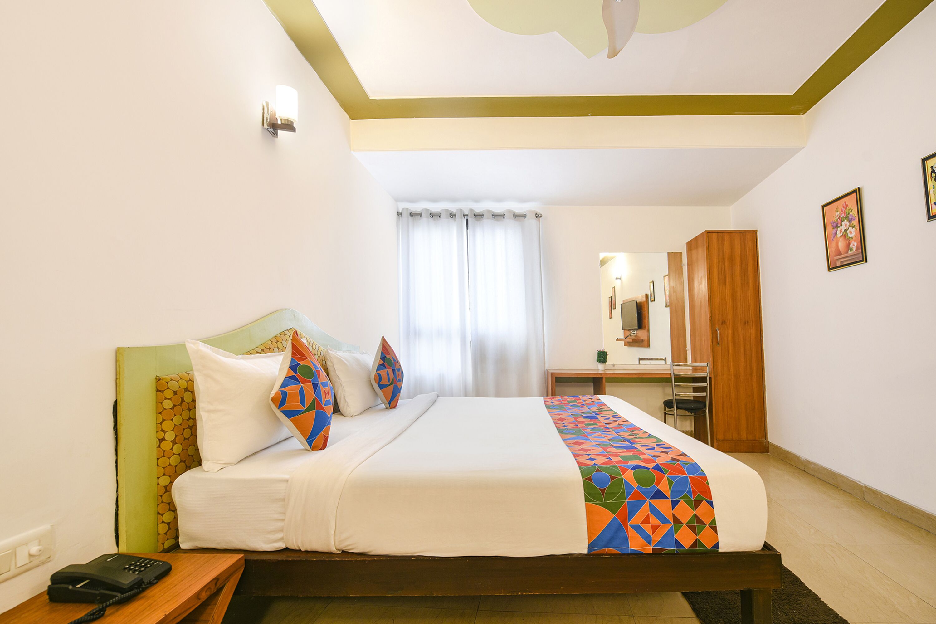 A hotel room with a bed, a bedside table with a phone, a wooden wardrobe, a mirror, and a window with curtains.