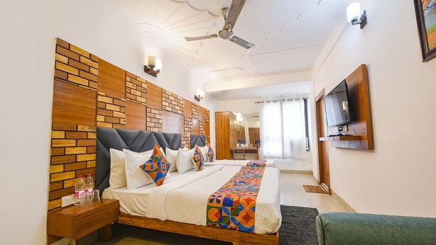Een hotelkamer met een houten hoofdeinde, een bed met een kleurrijke deken, een nachtkastje met flessen, een plafondventilator en een aan de muur bevestigde televisie.