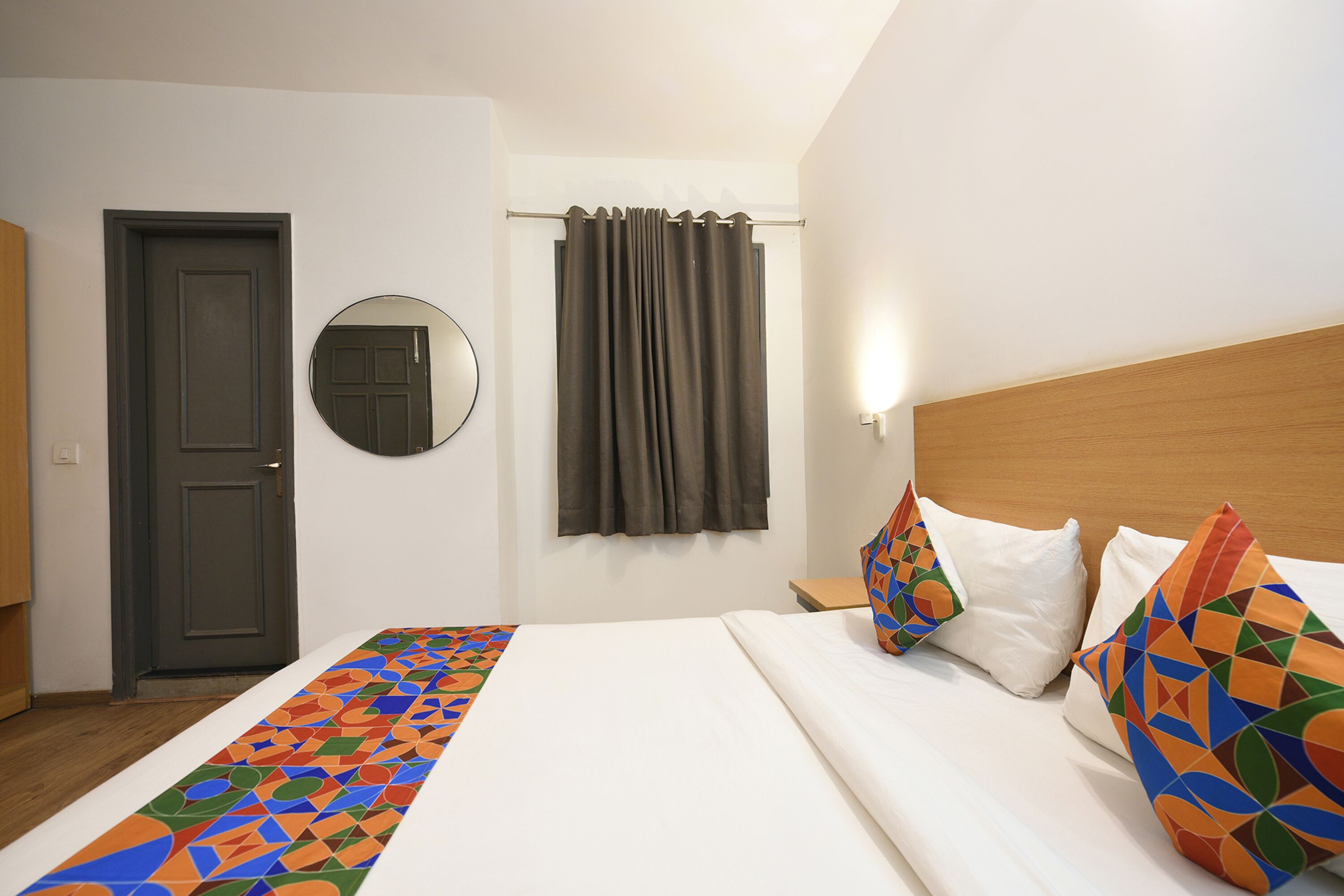 A hotel room with a bed, a wooden headboard, a mirror, a curtain, and a door.