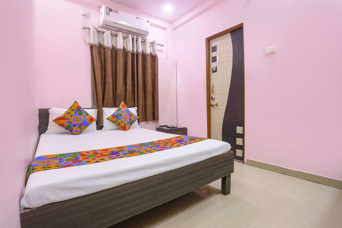 Una cama bien hecha con una colcha de diseño geométrico y llamativos colores, dos almohadas decorativas, una mesita de noche con una lámpara y un aire acondicionado instalado en la pared.