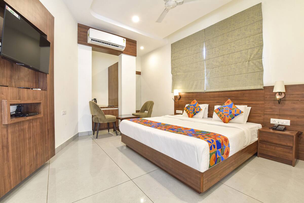 A hotel room with a large bed, a desk, a chair, an air conditioning unit, a television, and a bedside table with a lamp.