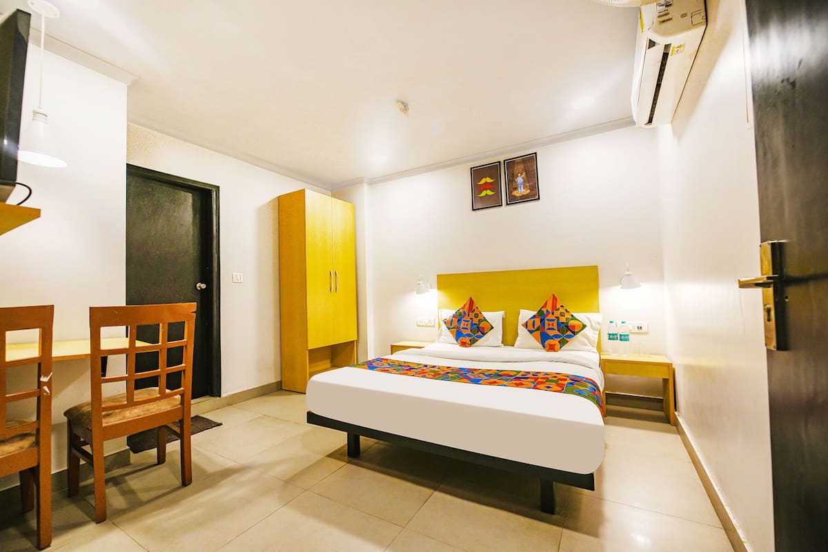 Habitación de hotel con una cama, un escritorio y una silla. Se observa una puerta, un aire acondicionado instalado en la pared y una mesita de noche con lámpara.