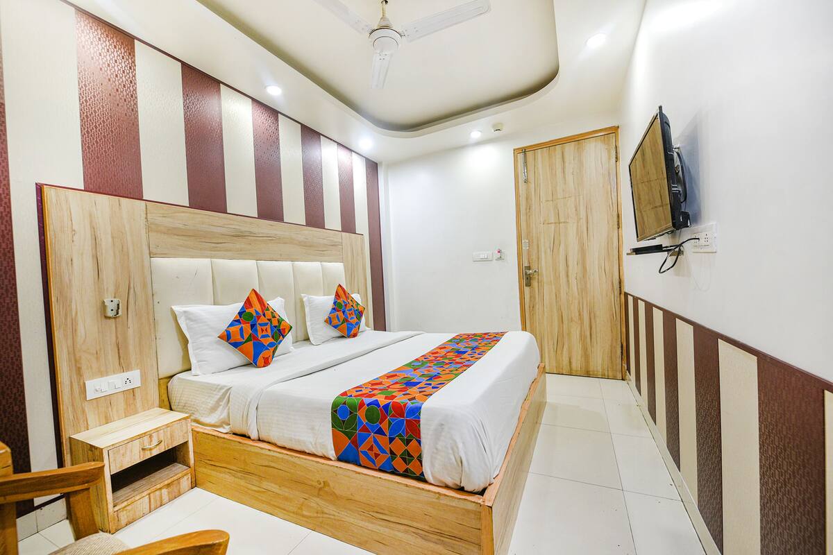 A hotel room with a wooden bed frame, a white headboard, and a colorful bedspread. There is a wooden nightstand with a drawer, a wooden chair, and a television mounted on the wall.