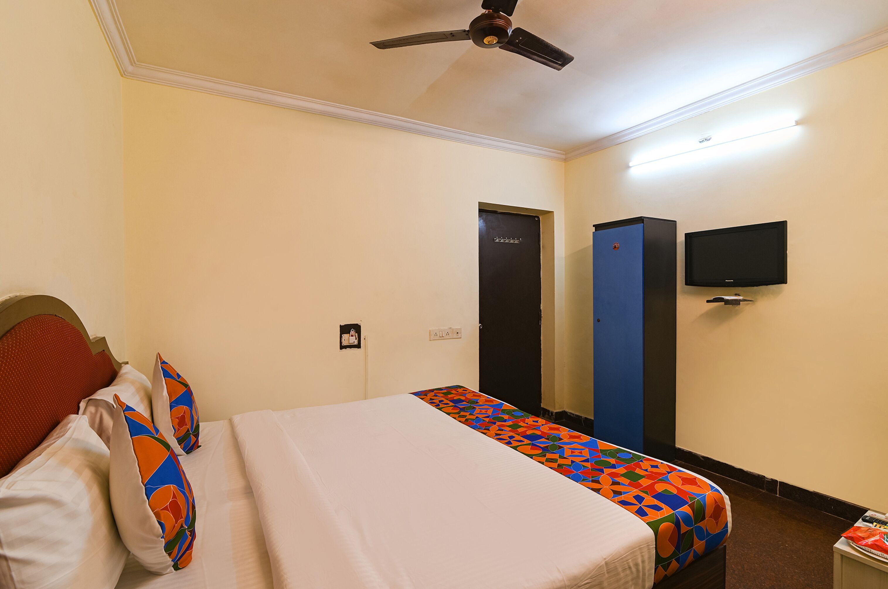 A hotel room with a bed, a ceiling fan, a TV mounted on the wall, and a door.