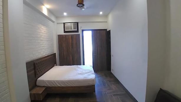 Un couloir étroit, avec un lit au fond, un banc en bois, un climatiseur fixé au mur et une porte avec un rideau.