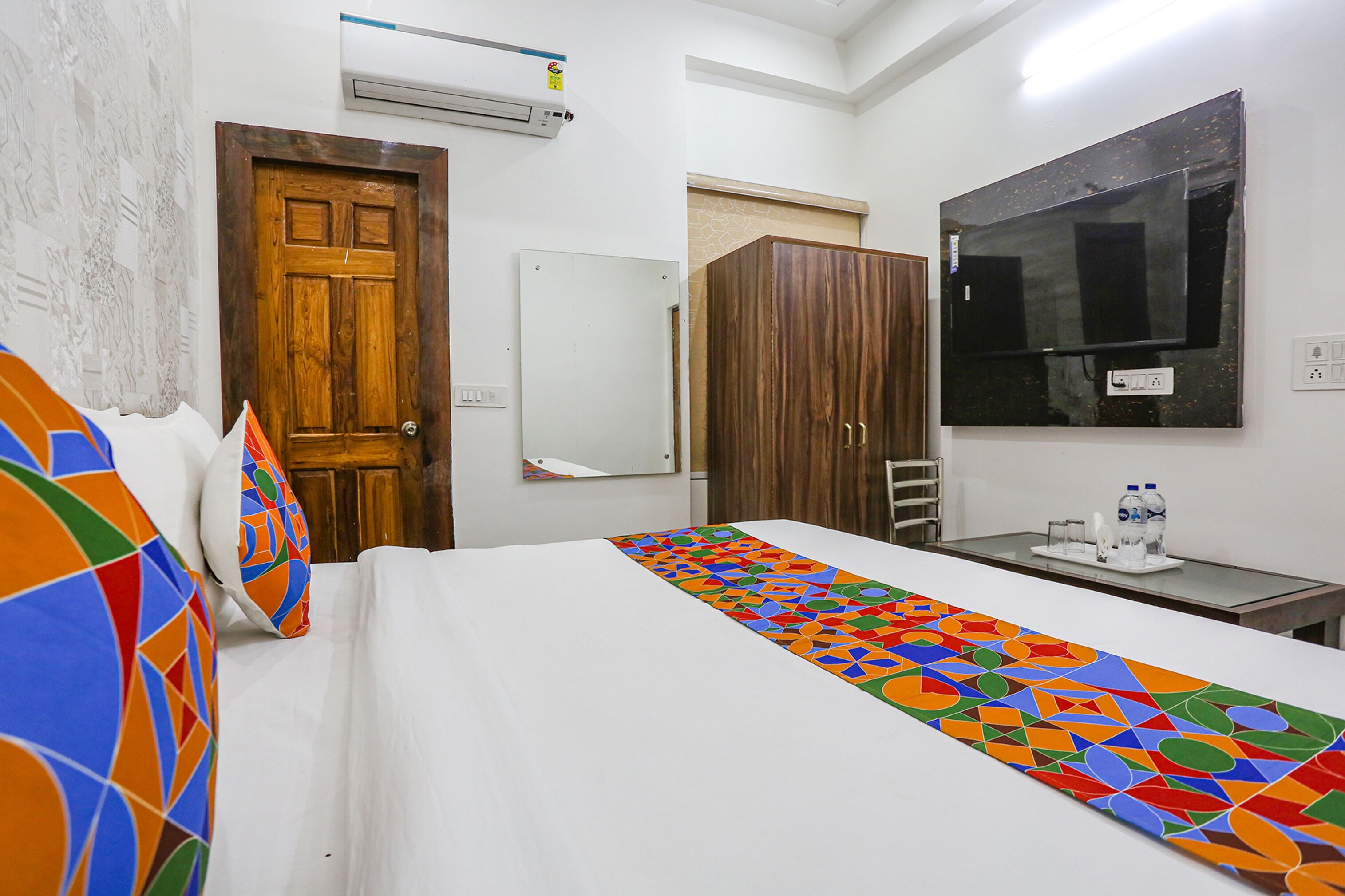 A hotel room with a bed, a wooden wardrobe, a mirror, a television, and a small table with a water bottle.