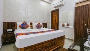 A bedroom with a wooden bed, white bedding, patterned pillows, a nightstand, a mirror, a wardrobe, and an air conditioning unit. - Fabhotel Sangita Dham (Mathura)