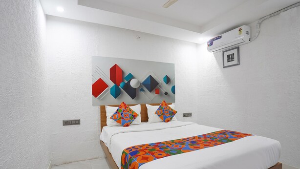 A bedroom with a bed, white walls, a ceiling fan, an air conditioning unit, and a geometric artwork above the bed. - FabExpress NK Comforts (Bengaluru)