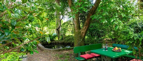 A stone-paved garden with a green picnic table and benches, surrounded by lush greenery and trees.