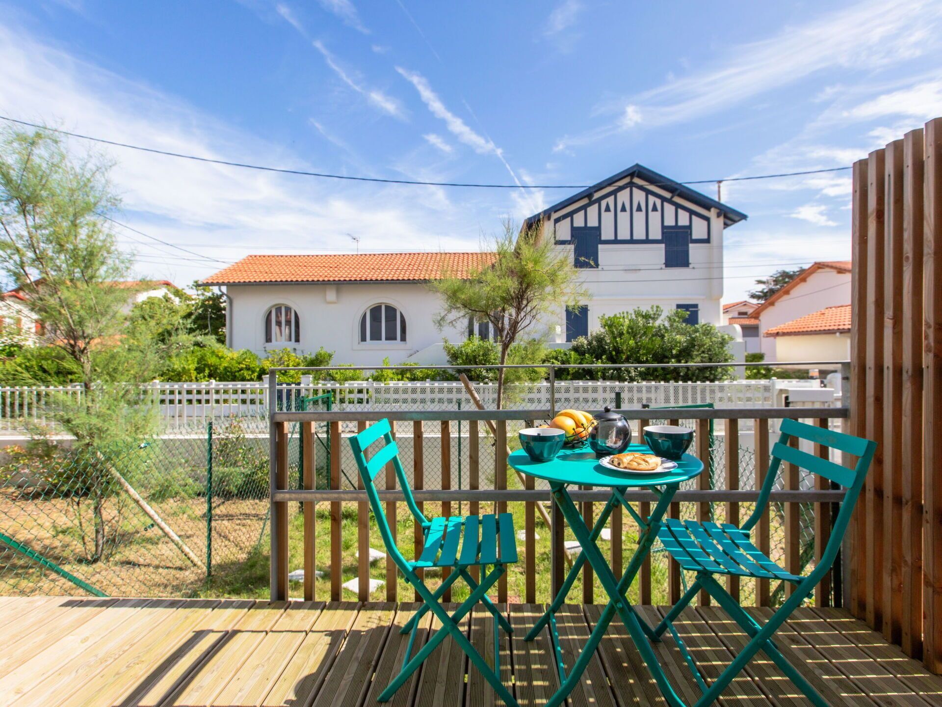 Eine Holzterrasse mit Tisch und Stühlen, ein weißes Gebäude mit rotem Dach im Hintergrund und ein weißer Zaun.