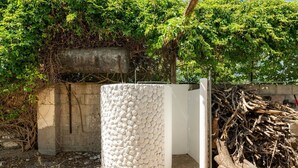 Eine Betonkonstruktion mit strukturierter Oberfläche, umgeben von einem Holzstoß und einer mit Kletterpflanzen bewachsenen Mauer.