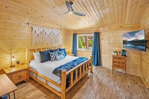 A wooden cabin bedroom with a bed, bedside table, lamp, and a TV on a dresser.