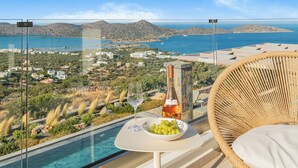 A balcony with a glass barrier, a wicker chair, a table with a bottle of wine, a glass, and a bowl of grapes.
