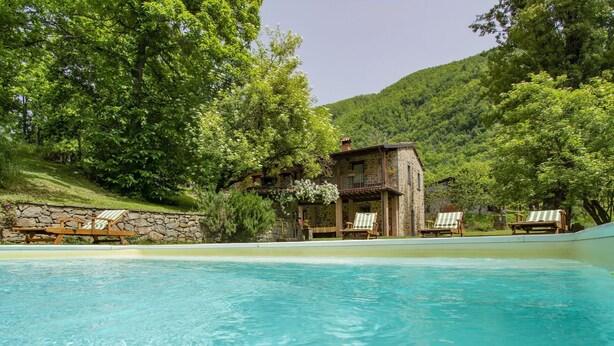 Ein Pool mit Liegestühlen, ein Steinhaus und üppiges Grün.