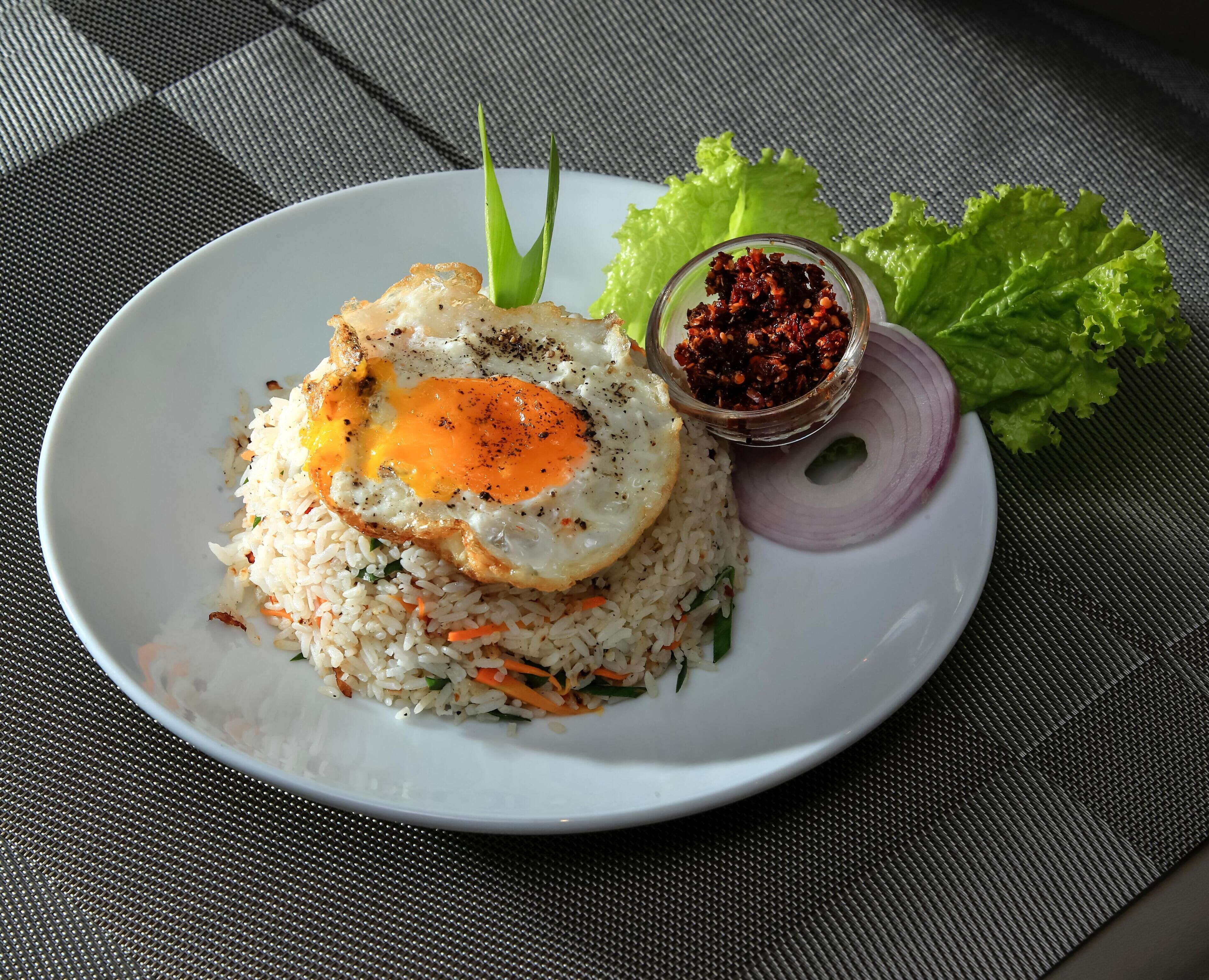Un plato de arroz frito coronado con un huevo estrellado, acompañado de un tazón pequeño de salsa de chile rojo, lechuga y una rodaja de cebolla roja.