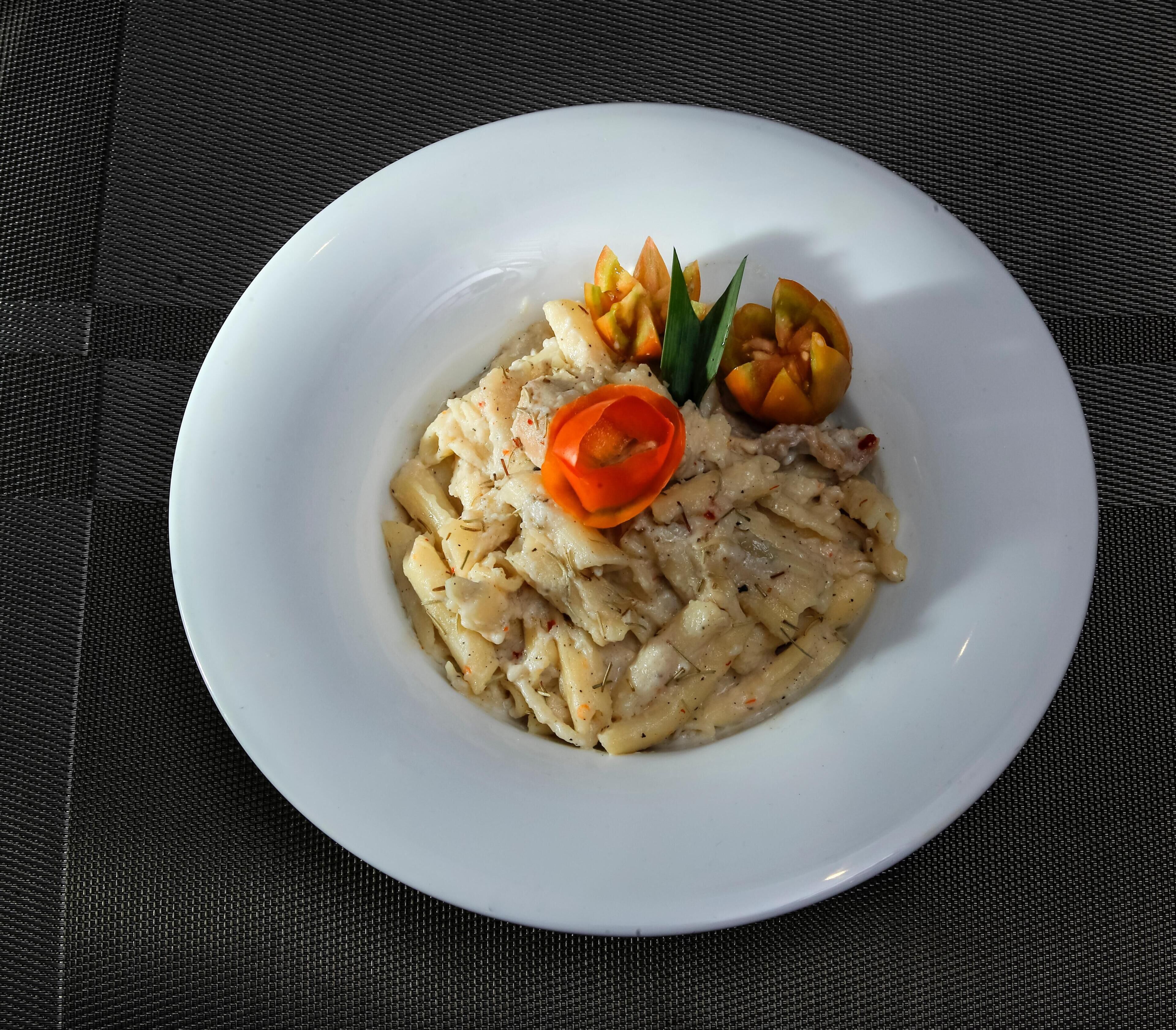 A white plate with pasta, garnished with a red chili and two orange slices, on a textured black surface.