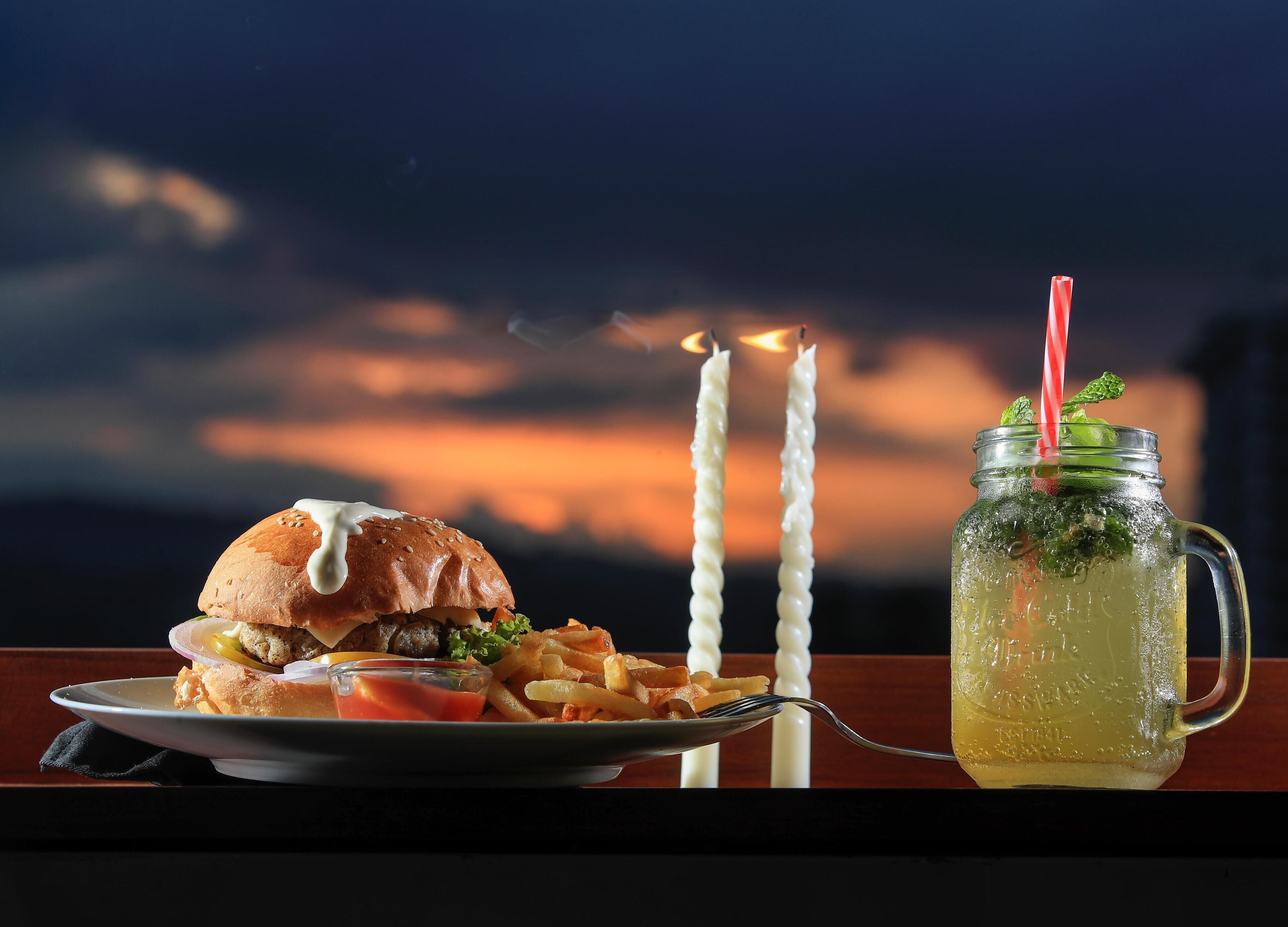 A meal with a burger, fries, and a drink in a mason jar, with candles in the background.