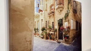 Un tableau représentant une rue étroite, bordée de bâtiments des deux côtés, avec des balcons ornés de plantes et un pavé.