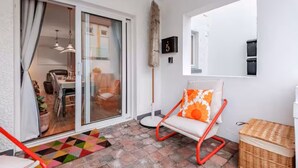 Ein Zimmer mit einer Schiebetür und Glasfront, die in einen Essbereich führt, ein roter Sessel mit orangefarbener Sitzkissen, ein kleiner Korbwäscher und ein Fliesenboden.