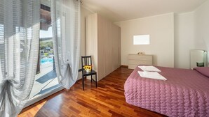 A bedroom with a wooden floor, a bed with a pink quilt, a chair with a vase of sunflowers, a dresser, and a window with sheer curtains.