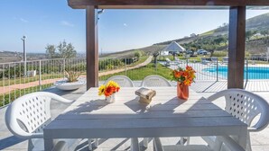A covered outdoor seating area with a table and chairs, overlooking a swimming pool and a scenic landscape.