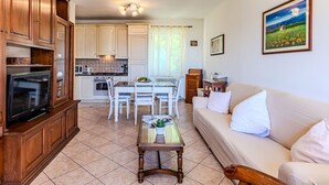 Een woonkamer met een bank, een salontafel en een televisie. Er is een keuken met witte kasten en een eethoek met een tafel en stoelen.