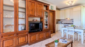 Een ruime woonkamer met houten kastjes, een flatscreen televisie en een eethoek met witte stoelen. Op de achtergrond is een keuken te zien.