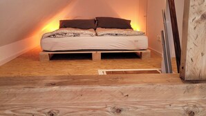 Une chambre située dans un espace de type « loft », avec un plafond en pente, un plancher en bois et un lit doté d’un matelas blanc et de draps à motifs.