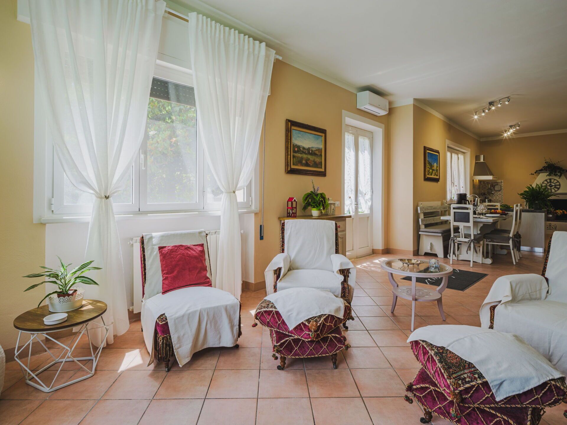 Un salón espacioso con zona de comedor, que cuenta con un ventanal con cortinas blancas, un cuadro en la pared y una cocina al fondo.