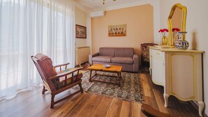 Una sala de estar con sofá, una mesa de centro de madera, alfombra estampada, una silla de madera, un espejo y una araña.