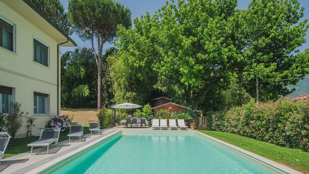 Piscina rectangular con agua cristalina, rodeada de césped y árboles. Se observan sillones de descanso y una zona de barbacoa al fondo.