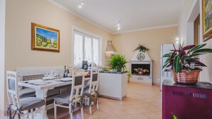 A dining area with a white table set for four, a coffee machine, and a window with curtains. There is a decorative plant on a stand and a clock on a mantelpiece.
