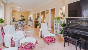 A spacious living room with a piano, white armchairs, and a dining area with a kitchen in the background.
