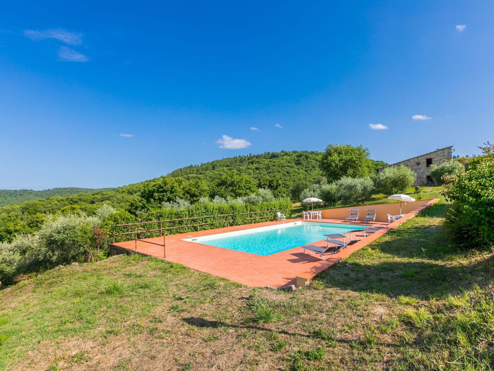 A rectangular swimming pool with a red border, surrounded by grass and trees, with a stone building in the background.