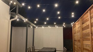 Un espace extérieur aménagé en terrasse, avec une table et des chaises en bois, des guirlandes lumineuses et une clôture en bois.