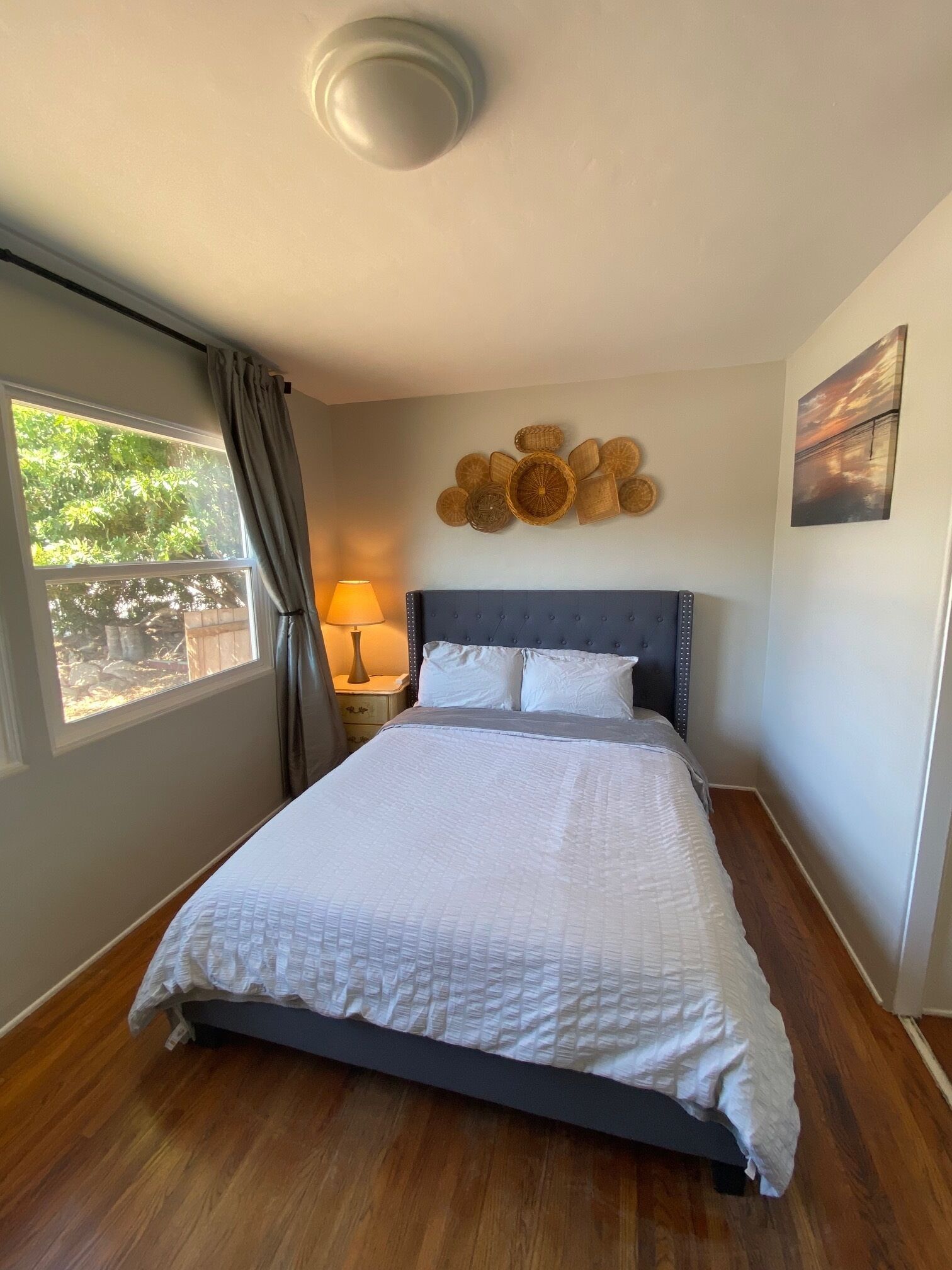 A bedroom with a bed, a nightstand, a lamp, a window with curtains, a wall art piece, and a ceiling light.