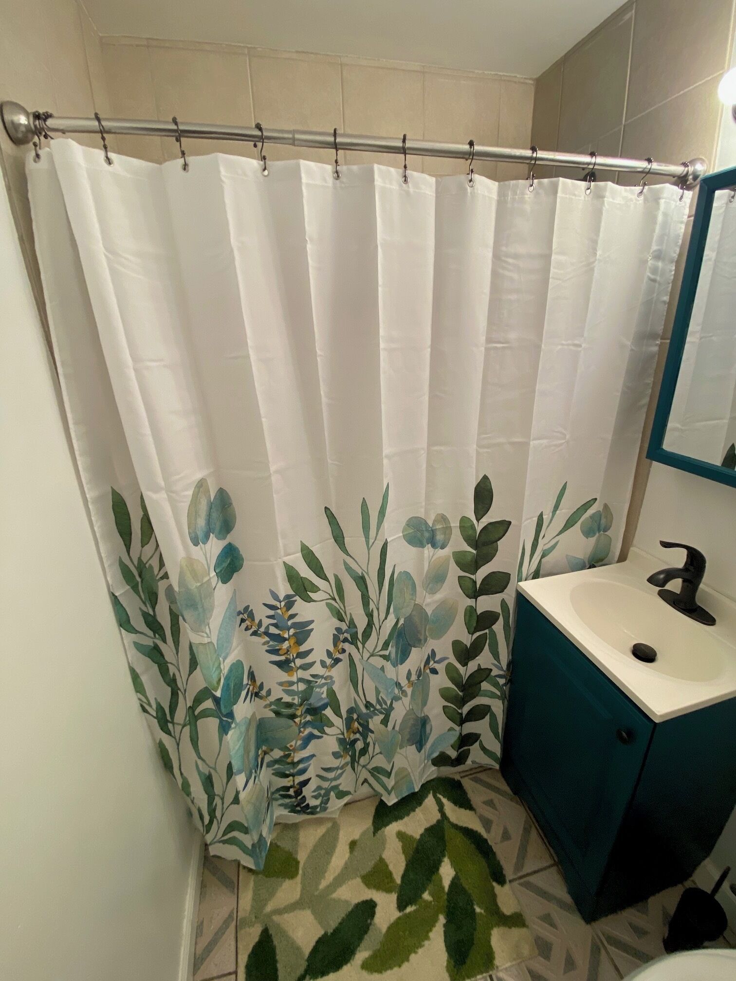 A bathroom with a shower curtain featuring a leaf pattern, a sink with a dark faucet, and a teal cabinet.