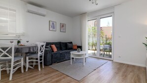 A modern living room with a dark grey sofa, white chairs, a small round table, and a glass door leading to a balcony with plants.